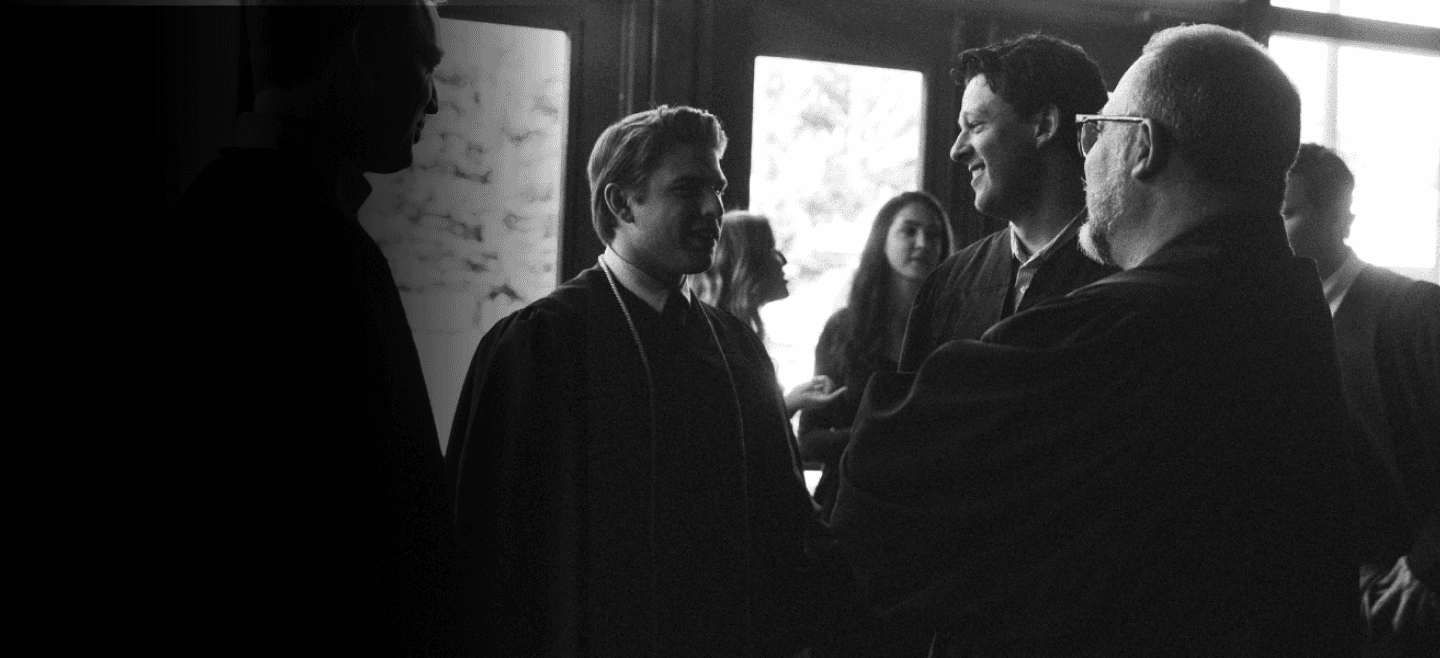 A college student smiling in graduation robes while talking to another graduating student and a professor
